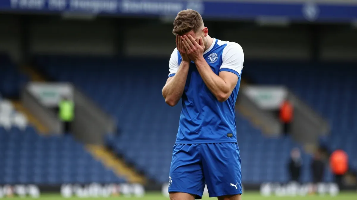 A Hartlepool United player, wearing a blue shirt with white sleeves, and blue shorts, with a white collar, looks...