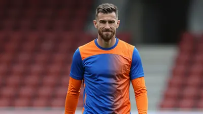 Mid-30s muscular striker, waist-up, wearing a plain Eastleigh home training top split blue left side, orange right side...