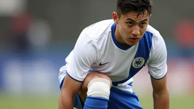A 21-year-old left-back, wearing a white jersey with a blue stripe, a blue short, and blue socks, with a blue and white...